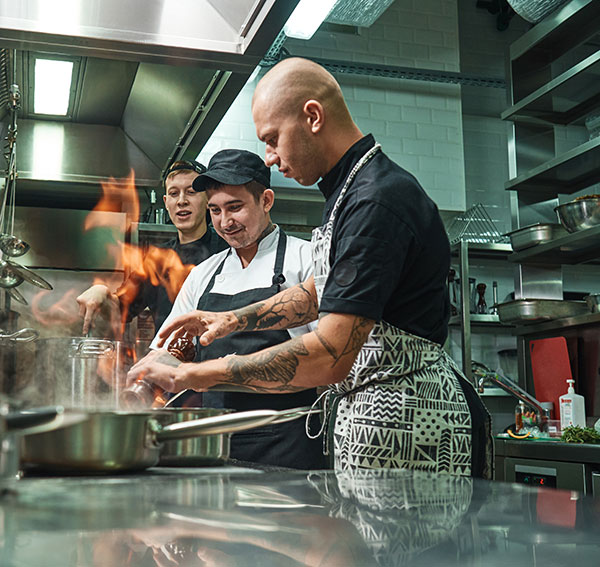 Two young students watch a man with tattoos preparing food