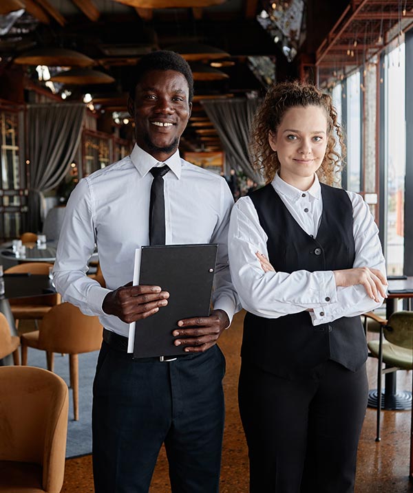 Trainee front of house staff, one black man and one white woman smile at camera